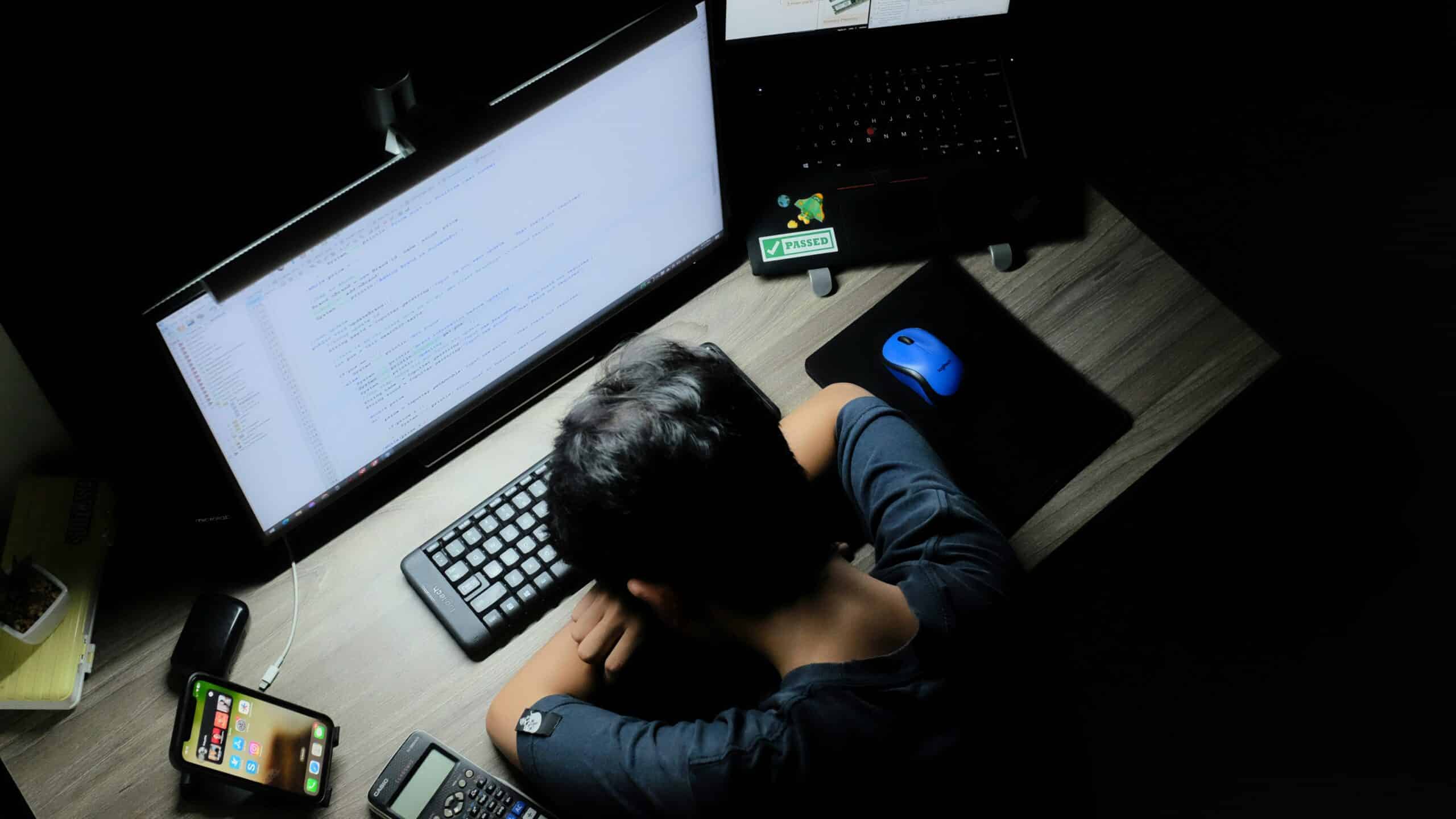 Overhead view of a person coding at a desk with a large monitor, laptop, keyboard, mouse, and phone in a dimly lit workspace.